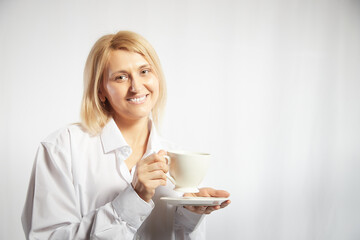Portrait of pretty blonde smiling woman posing with cup of tea or coffee on white background. Happy girl model in white shirt in studio. The concept of pleasant morning at home or at work. Copy space