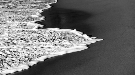Fototapety  Black sandy beach with splashing water, surf, bubbles, wave foam and low evening sunlight. Black and white greyscale. “Anse Couleuvre“ is a remote secluded beach on tropical island Martinique. 