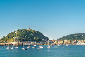 Naklejka premium SAN SEBASTIAN, Spain July 08 2022: View of Santa Clara Island. Boats docked in the middle of La Concha Bay. Beautiful travel destination in north of Spain. Jesus Christ Statue on top of the hill.