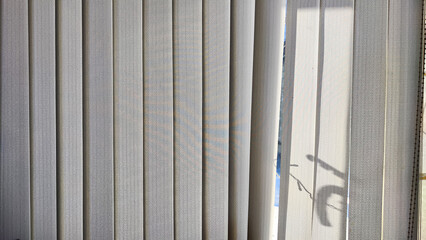 Obraz premium Vertical fabric white blinds on the window with light and shadow from the sun. Background and texture. Abstract pattern and frame. Stripes and lines of white-gray color