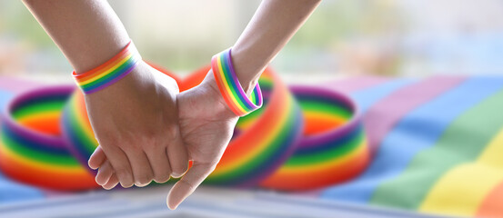 Closeup view of hands of gay couple wearing rainbow wristbands to present LGBT love, blurred rainbow flags background, concept for LGBT people celebrations in pride month.