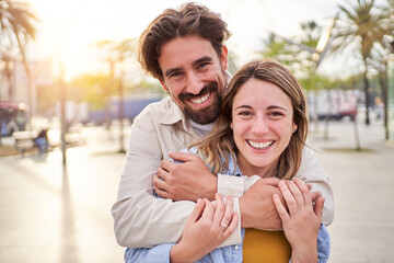 Caucasian couple looking at camera cheerfully while standing outdoors together. Husband embracing wife from behind with love during beach trip. Copy space.