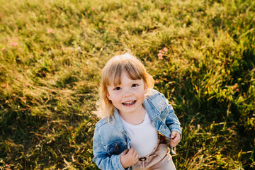 Top view on smiley blonde caucasian little girl in casual running on meadow looks atrocious camera happily. Cheerful Italian toddler enjoying sunset time outdoors. Nature, happy kids.
