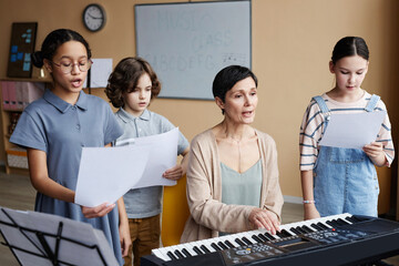 Group of children singing songs together with teacher while she playing piano during music lesson