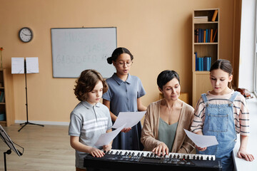 Group of children reading notes and singing songs while teacher playing piano during music lesson...