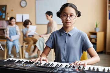 Portrait of schoolgirl in eyeglasses looking at camera while playing piano in music class with children and teacher in background
