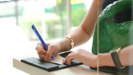 A woman signs a bill at a checkout counter after shopping. She looks at the bill and confirms the total amount before signing. The cashier hands her a shopping bag with her purchased items, and she