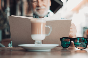 Blurred senior man sitting outdoor at a cafe table reading a book while has a break with a coffee and milk drink - caucasian elderly man relaxed in retirement or vacation