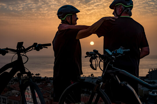 Two senior cyclists in silhouette in sunset light with ocean as backdrop standing near bicycles - healthy lifestyle in retirement concept