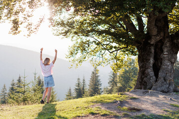 A charming picturesque view of the mountains. A young woman is standing near a big tree on a hill and raised her hands up. Carpathian Mountains