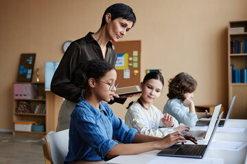 Group of school children using computers during study with teacher helping them with online presentation