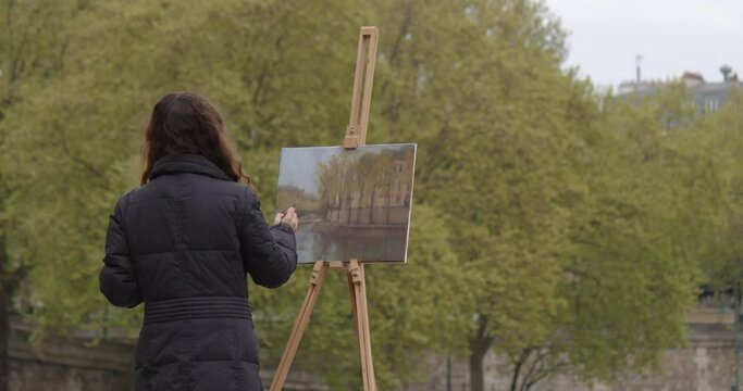 Woman painting a picture near the river Seine. Creative woman artist paints a picture with a brush while standing near the easel in Paris. Woman artist, art for sales, inspiration