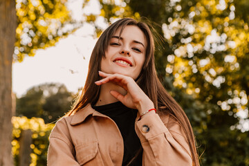 Caucasian young pretty stylish woman standing outside street dressed brown trench coat smiling poses outside city, spring autumn season nature sunshine light leaks. Cute brunette hair lady 