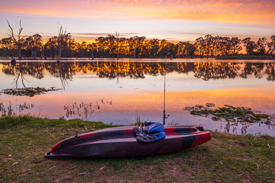 A fishing kayak sitting on the edge of a river under a dramatic sunrise