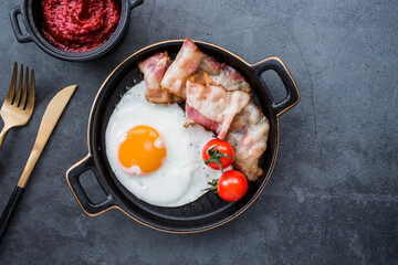 Fried eggs on a stylish ceramic plate with handles with fried bacon, cherry tomatoes and tomato paste on a dark background. Breakfast, lunch and dinner. Delicious food. Cooking.