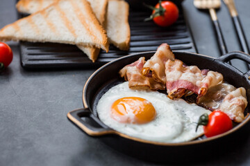 Fried eggs on a stylish ceramic plate with handles with fried bacon, cherry tomatoes, tomato paste and bread toast on a dark background. Breakfast, lunch and dinner. Delicious food.