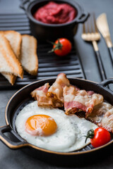 Fried eggs on a stylish ceramic plate with handles with fried bacon, cherry tomatoes, tomato paste and bread toast on a dark background. Breakfast, lunch and dinner. Delicious food.