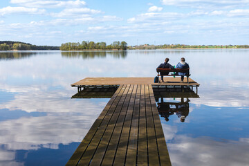 A pair of men, father and son, sitting on a wooden dock by the lake and admiring the view of the water and blue sky