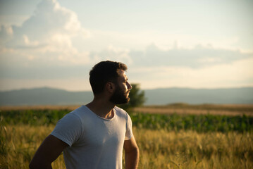 Naklejka na ściany i meble Happy young handsome man spending time in nature during the golden hour. Enjoying in sunset and looking to the sun. Walking through countryside.