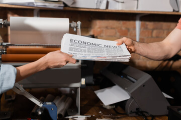cropped view of typographer giving newspaper with economic news lettering to colleague in print center.