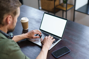 man working on laptop in loft office. man seated at desk with laptop, mobile phone, and coffee paper cup placed on it. typical workspace setup, giving viewer sense of modern work environment.