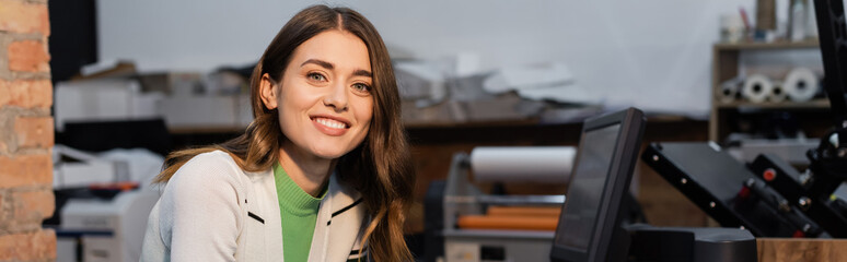 cheerful woman smiling near monitor while working in print center, banner.