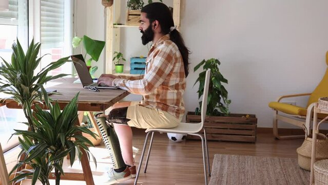 millennial man with long hair and beard, telecommuting work at home, has amputated leg wears a prosthesis.