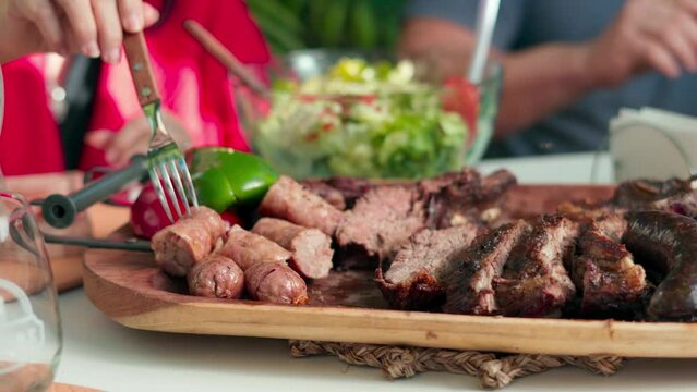 closeup of several people's hands holding forks while skewering pieces of roast meat served on a wooden board on a table with a bowl of salad outdoors during daytime