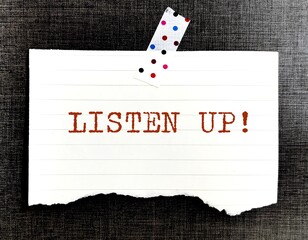 Stick note on wallpaper with handwritten text LISTEN UP!, means to tell others to listen closely to...