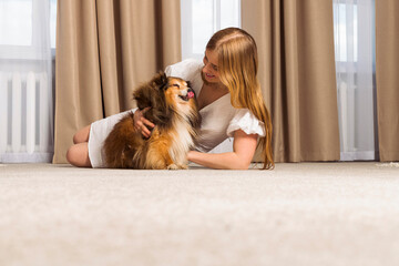 Adorable aged Miniature Sheltie dog and its owner, cute blonde girl are spending time together indoors. Strong friendship and funny leisure with the pet.