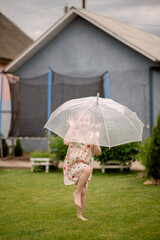 A cheerful little girl is holding a protective umbrella, feeling protected and full of happiness on a rainy day of her childhood. The child is having fun in the backyard of her house.