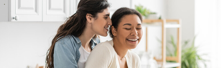 cheerful lesbian woman hugging multiracial girlfriend in modern kitchen, banner.