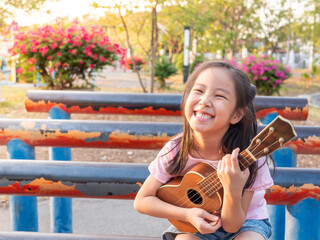 Little asian child girl play the ukulele, in the garden on the Steel pipe