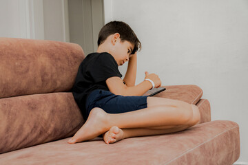 The boy plays on the sofa in the living room. Living room kitchen with sofa. The child plays with gadgets. The boy spends his summer vacation playing games on his phone.