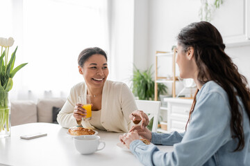 cheerful multiracial lesbian couple smiling while having breakfast in kitchen.