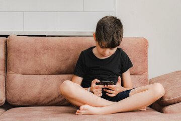 The boy plays on the sofa in the living room. Living room kitchen with sofa. The child plays with gadgets. The boy spends his summer vacation playing games on his phone.