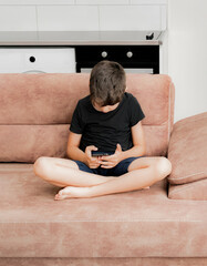 The boy plays on the sofa in the living room. Living room kitchen with sofa. The child plays with gadgets. The boy spends his summer vacation playing games on his phone.