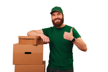Young handsome delivery man is holding thumb up while leaning on some boxes over white background.