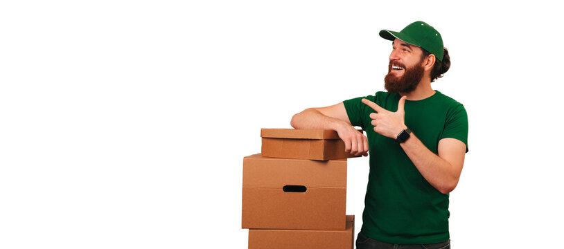 Cheerful young bearded man is pointing aside while leaning over some boxes. Banner shot over white background.