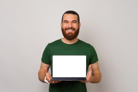 Wide smiling bearded man is holding a laptop and showing blank screen over grey background,.