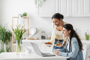 happy multiracial woman holding plates with croissants near girlfriend working on laptop from home.