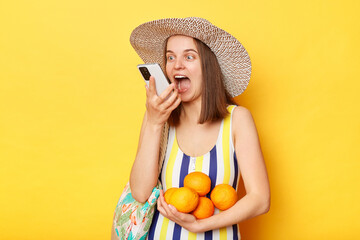 Happy woman wearing striped swimsuit and straw hat isolated yellow background using smart phone recording voice message on summer vacation.