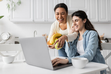 joyful multiracial woman holding bowl with fruits near girlfriend working on laptop at home.