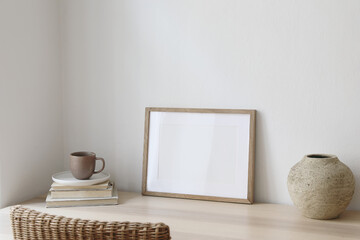 Empty horizontal picture frame mockup in minimalist interior. Wooden table, blurred rattan chair....