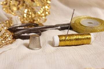 Fototapeta na wymiar Spool of Gold Thread and Scissors With Thimble on Off White Fabric With Gold Sequins