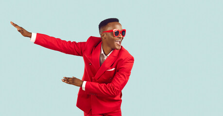 Happy young man in stylish party outfit dancing and having fun in studio. Cheerful excited African American man in red suit and sunglasses dancing isolated on light blue background. Fashion concept - Powered by Adobe