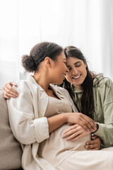 cheerful lesbian woman hugging pregnant multiracial wife while sitting on sofa.
