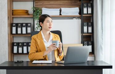 Asian businesswoman sits in a office working on laptop and enjoys a coffee.