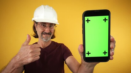 Mature elderly construction man wear hard hat point finger on mobile cell phone with blank screen workspace area tracking points on solid yellow background studio portrait.