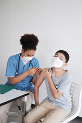 Fototapeta premium Professional African doctor or nurse woman is vaccinating to Caucasian boy is wearing mask in the hospital. Vaccines for children to prevent COVID 19 in hospital